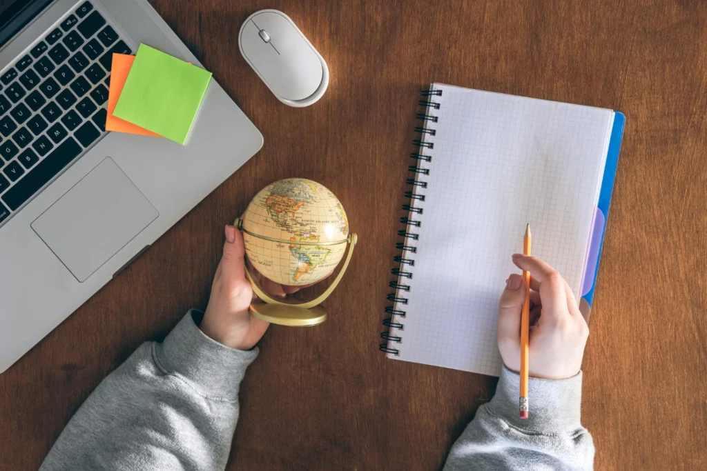 Hands holding a small globe next to a laptop, mouse, and notebook on a wooden desk, illustrating international trademark filing and global business protection.
