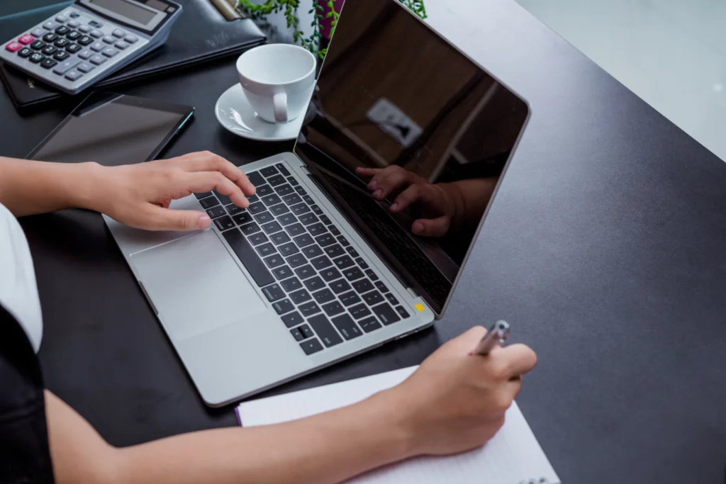 Close-up of a business owner working on a laptop and taking notes at a desk, representing the continuous commercial operations required for genuine use of a trademark.