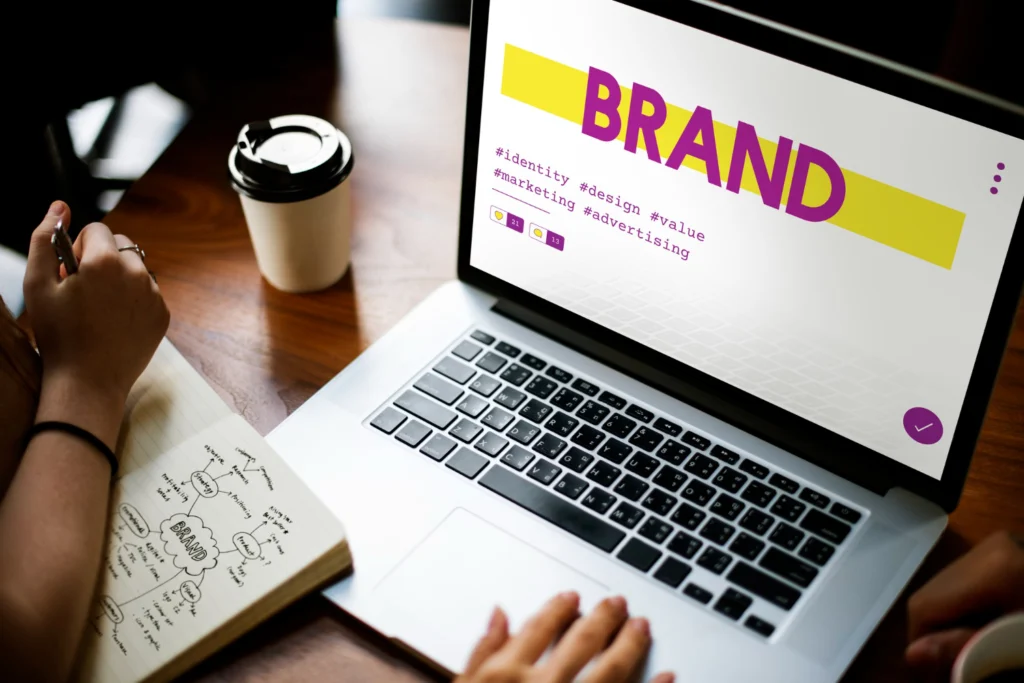 A laptop displaying a branding presentation next to a hand writing notes in a sketchbook, illustrating the active management and protection of trademarked brand assets.