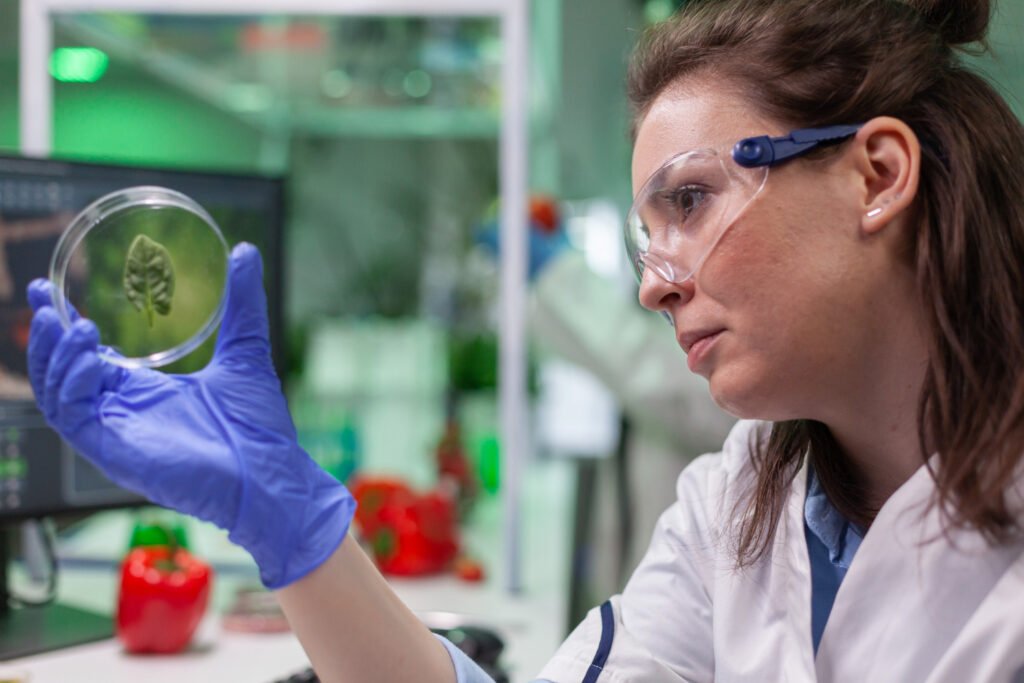 Female scientist in safety glasses examining a plant sample in a petri dish in a laboratory, with text about Microorganism Patent India deposit and disclosure rules.